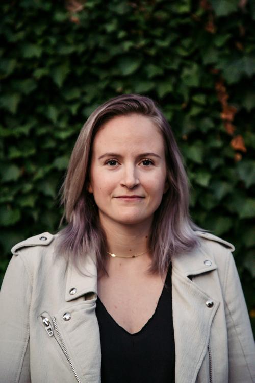 Woman with pale purple hair in light grey motorcycle jacket standing in front of a green wall of ivy.