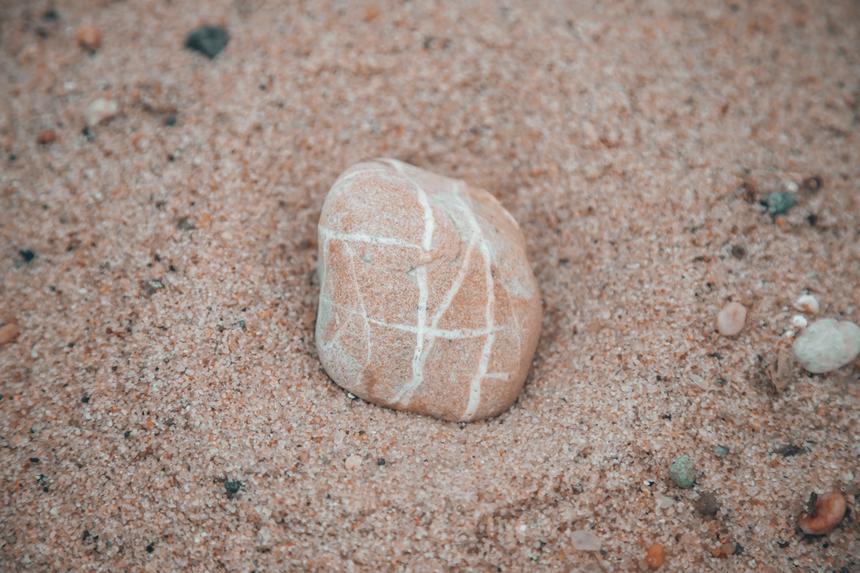 A unique rock with naturally occurring line markings that is sitting surrounded by sand.