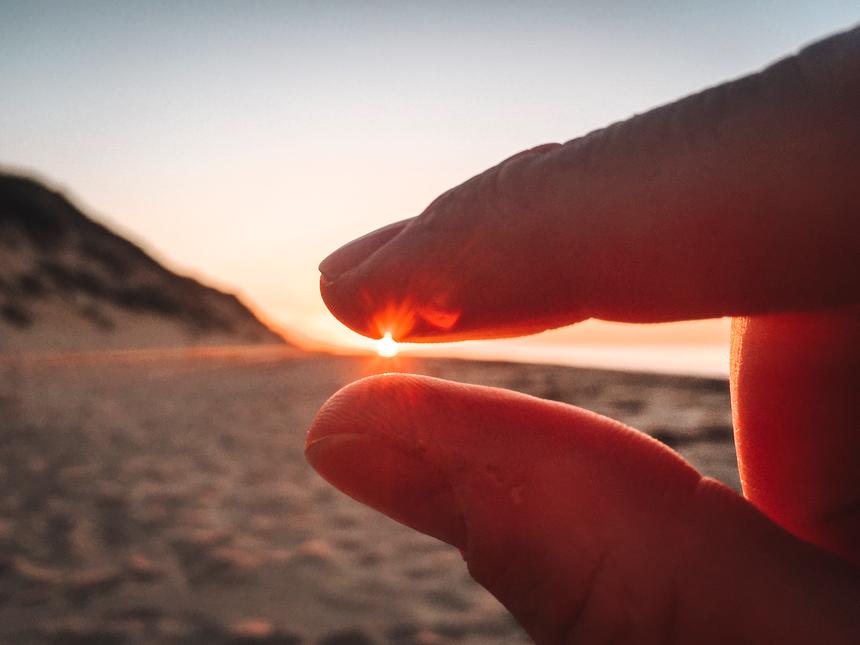 An up-close view of a hand with the finger and thumb closing around the sun setting in the distance.