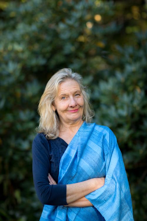 Woman with blue shawl draped over one shoulder who is smiling while standing in front of green plants