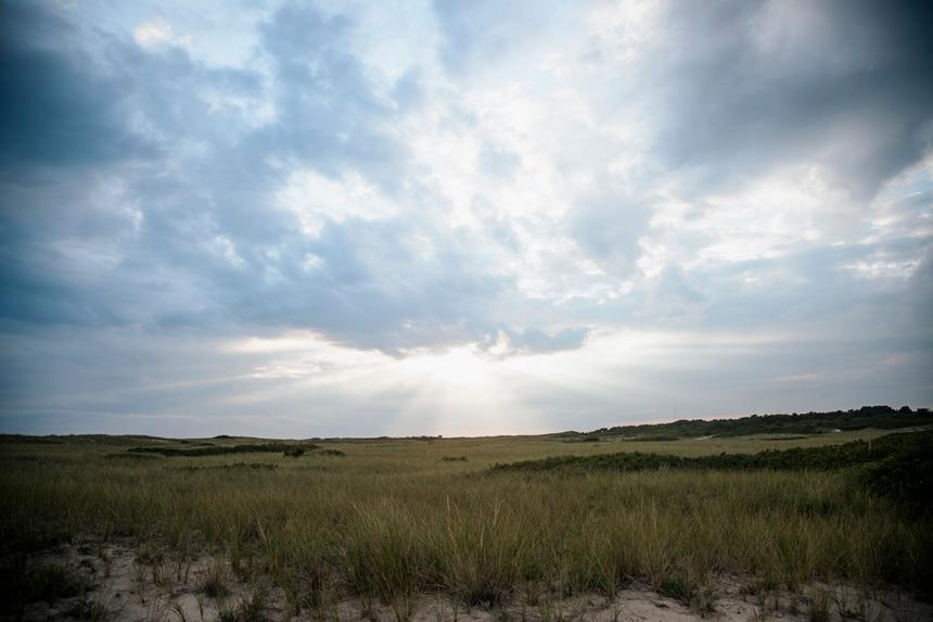 A landscape of cloudy skies with light breaking though over dune grasses.