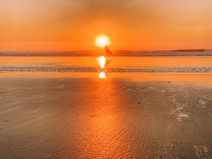 A surfer walks at the edge of the water with their surfboard with the sun setting behind them.